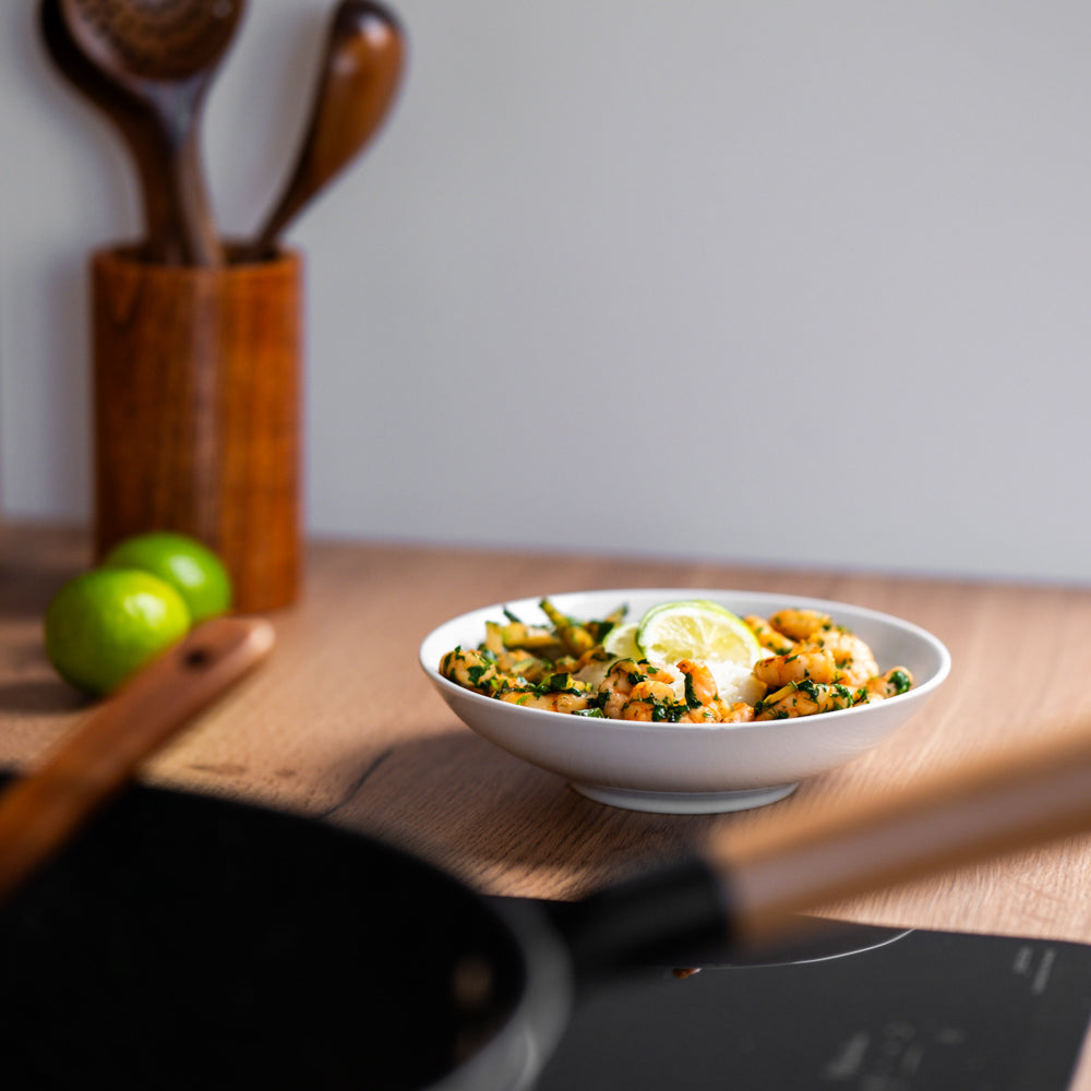 Skål med rejer, grønne krydderurter og lime på et køkkenbord, med wokens skaft uskarpt i forgrunden