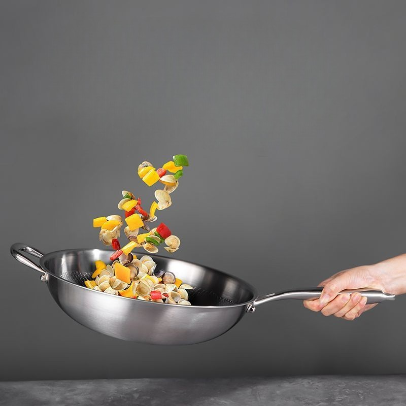 En hånd holder woken i skaftet og kaster muslinger og peberfrugt op i luften foran en grå væg
