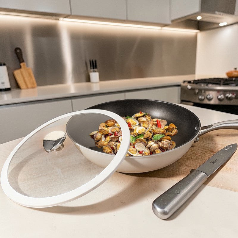 Hvid wokpande med muslinger, chili og krydderurter, glaslåget lænet op ad siden og en kniv på skærebrættet