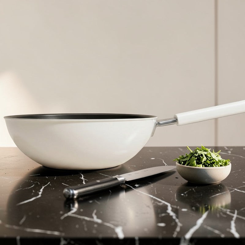 Hvid wokpande uden låg på sort marmorbord ved siden af en kniv og en lille skål hakkede krydderurter