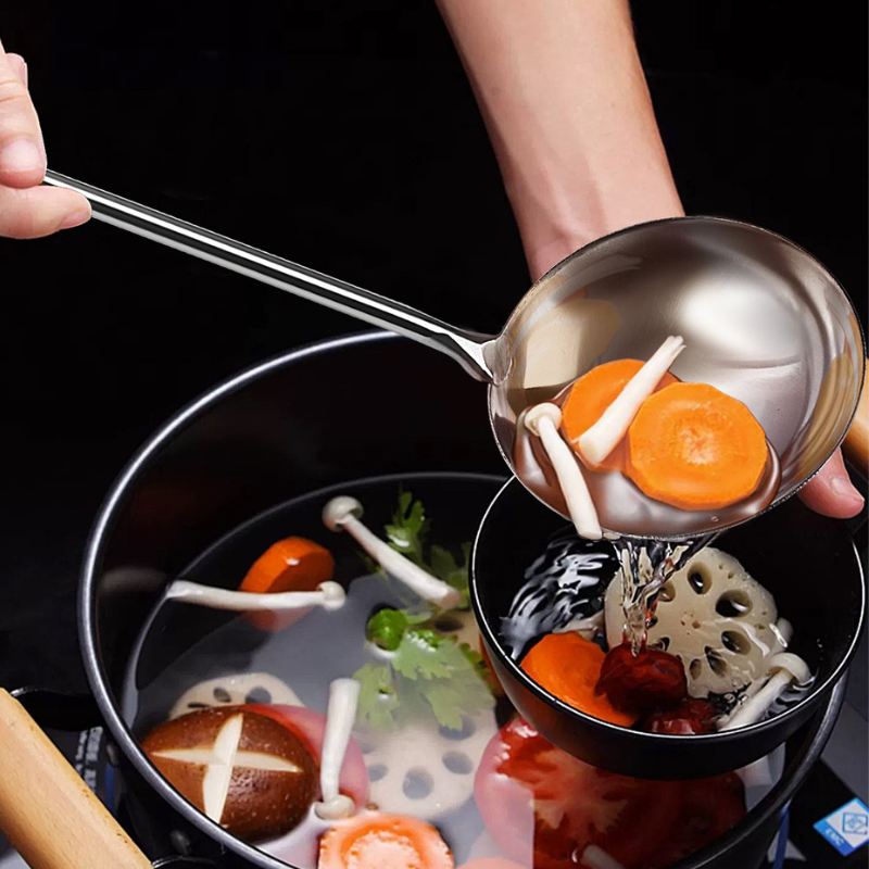 En hånd øser klar bouillon med gulerod og svampe fra en gryde over i en sort skål med wokskeen