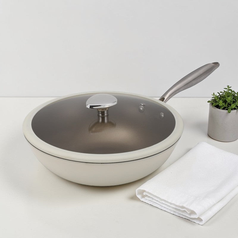 Hvid wokpande med glaslåg på et lyst bord ved siden af et foldet viskestykke og en lille potteplante