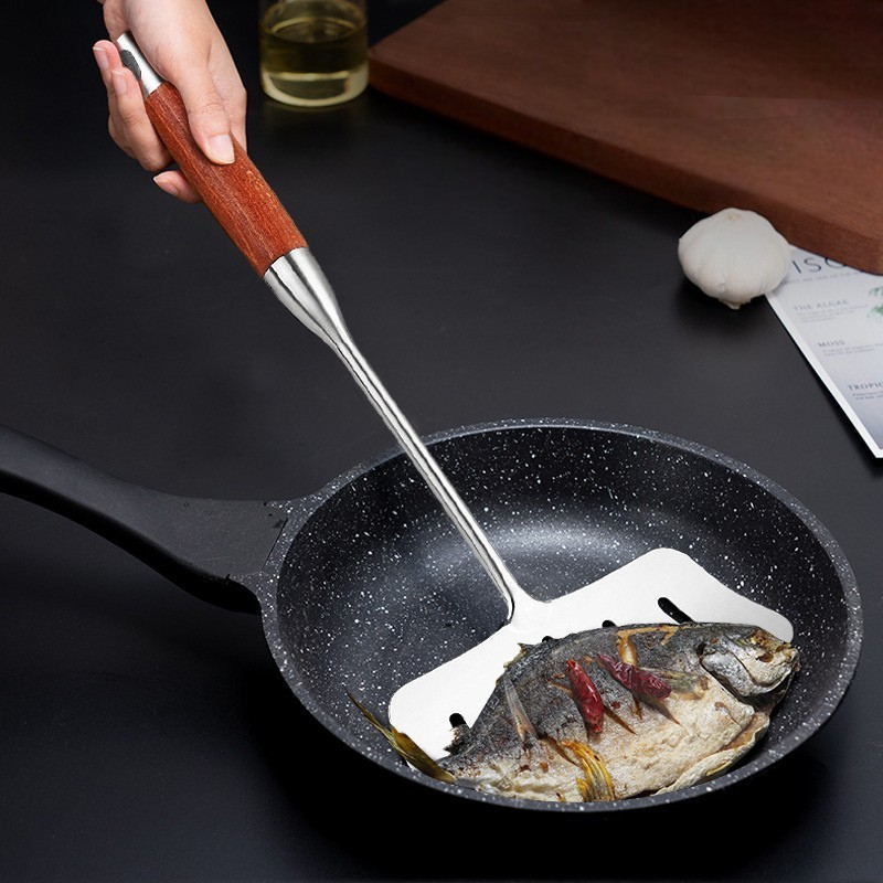 Hånd løfter en hel stegt fisk med tørret chili op af en sort stegepande med den brede palet