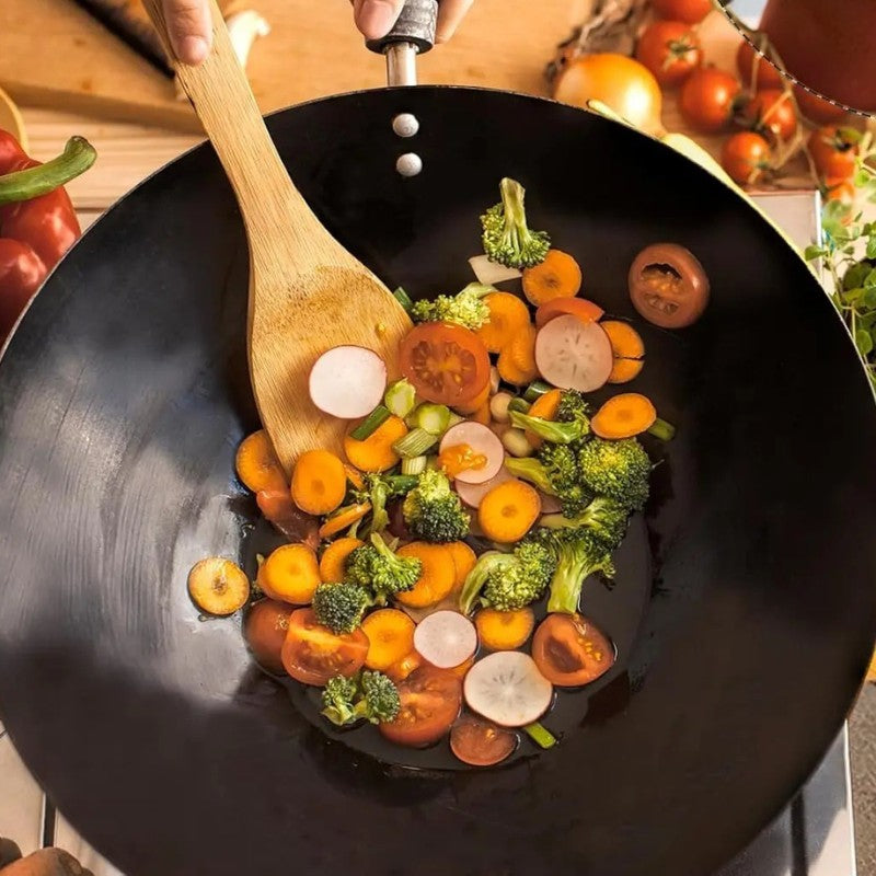 En hånd holder skaftet og vender gulerødder, broccoli og radiser med en træspatel, set oppefra