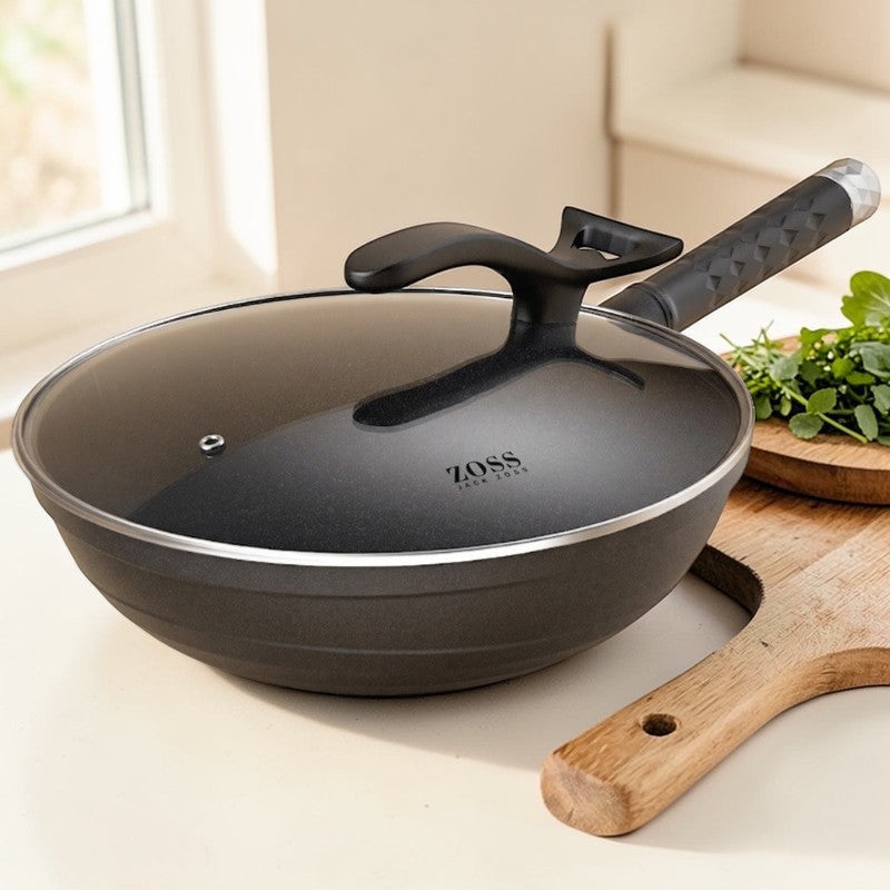 Sort wok med glaslåg på et lyst køkkenbord ved siden af et skærebræt i træ med friske krydderurter