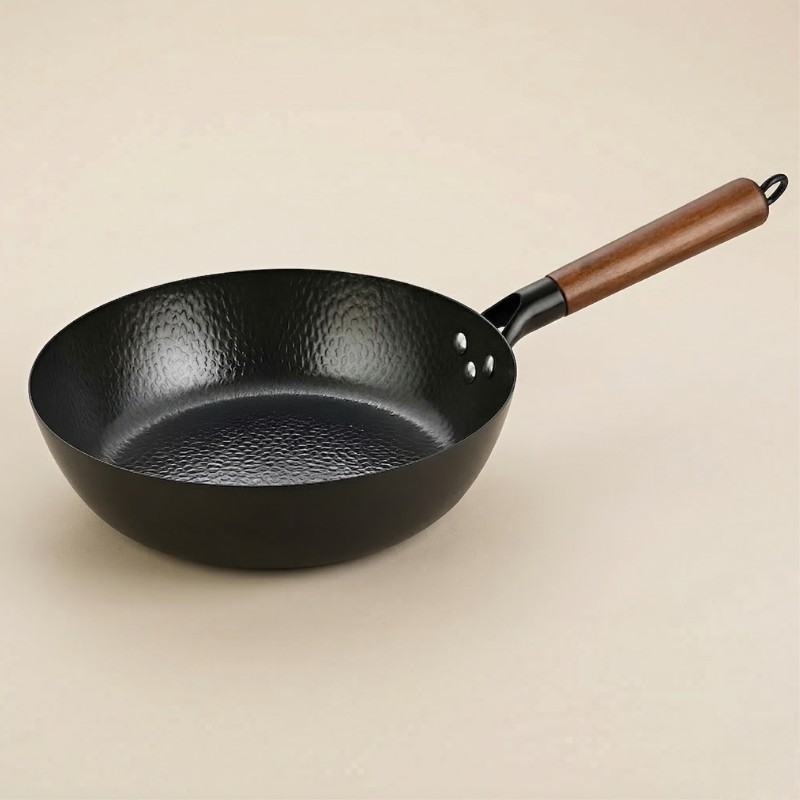 Le Rokko kulstofstål wok med sort hamret overflade og træhåndtag med ophængsring, set skråt fra oven på lys baggrund