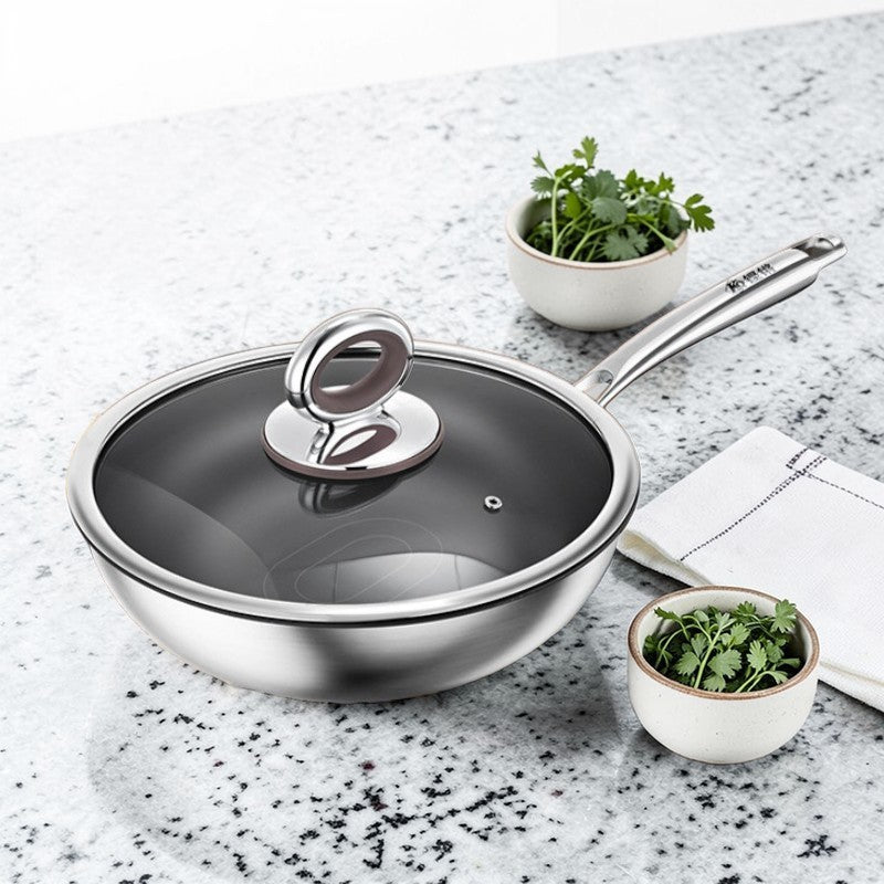 Wok i rustfrit stål med glaslåg på en spættet stenbordplade ved to skåle med koriander og en hvid serviet