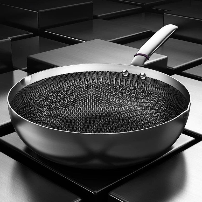 Wok i rustfrit stål uden låg på mørk baggrund, set skråt ovenfra med bikubemønster indeni og to nitter ved skaftet