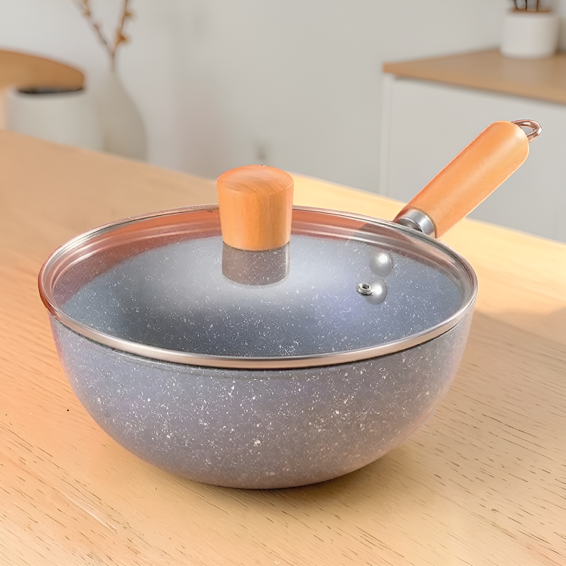 Mini wok med glaslåg på et køkkenbord i lyst træ, med et sløret køkken i baggrunden