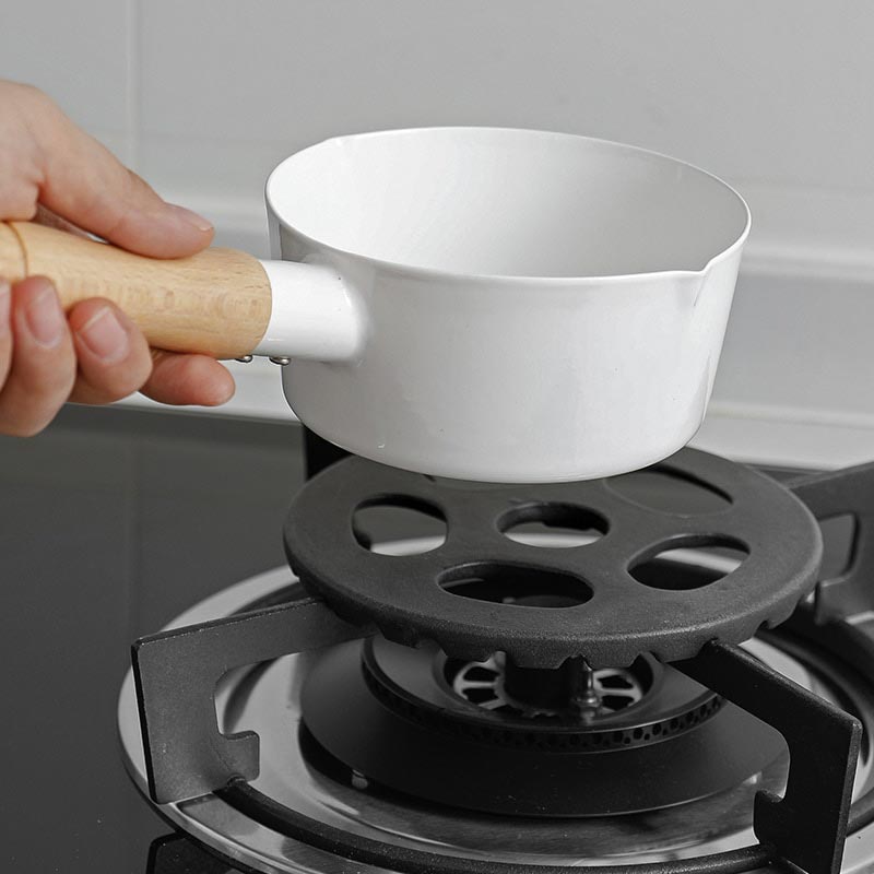 Hånd sætter en hvid kasserolle med træskaft ned på en rist til gasblus, der ligger oven på gaskomfurets rist