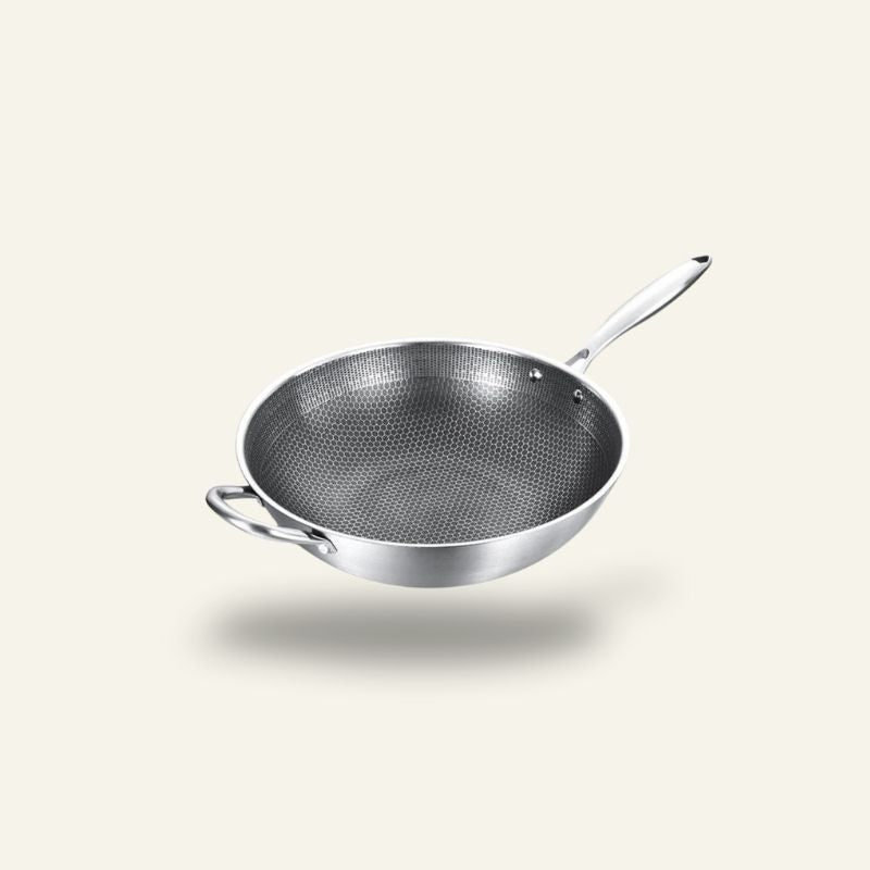 Le Kita wok i rustfrit stål med mørkt bikubemønster indeni, langt skaft og lille hank, svævende over en lys baggrund