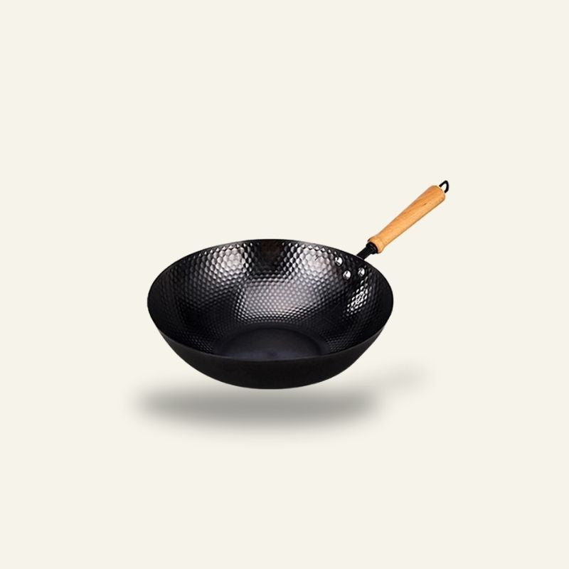 Le Kumichi wokpande i kulstål med mønstret inderside og langt håndtag af træ, set fra siden på lys baggrund
