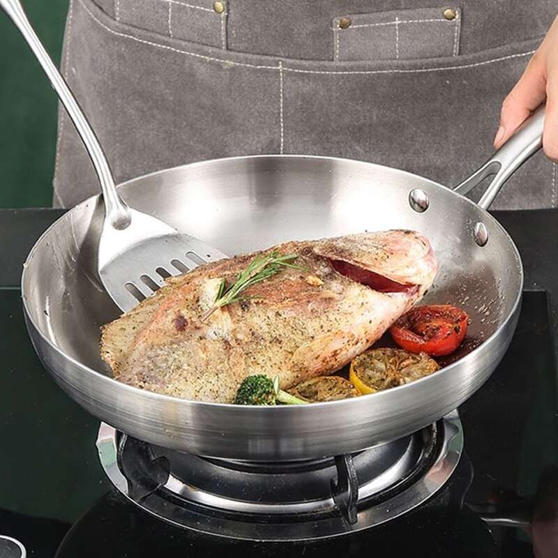 Stegt fisk med krydderurter og tomatskiver i panden på et gasblus, en hånd holder skaftet og en stegespatel i stål